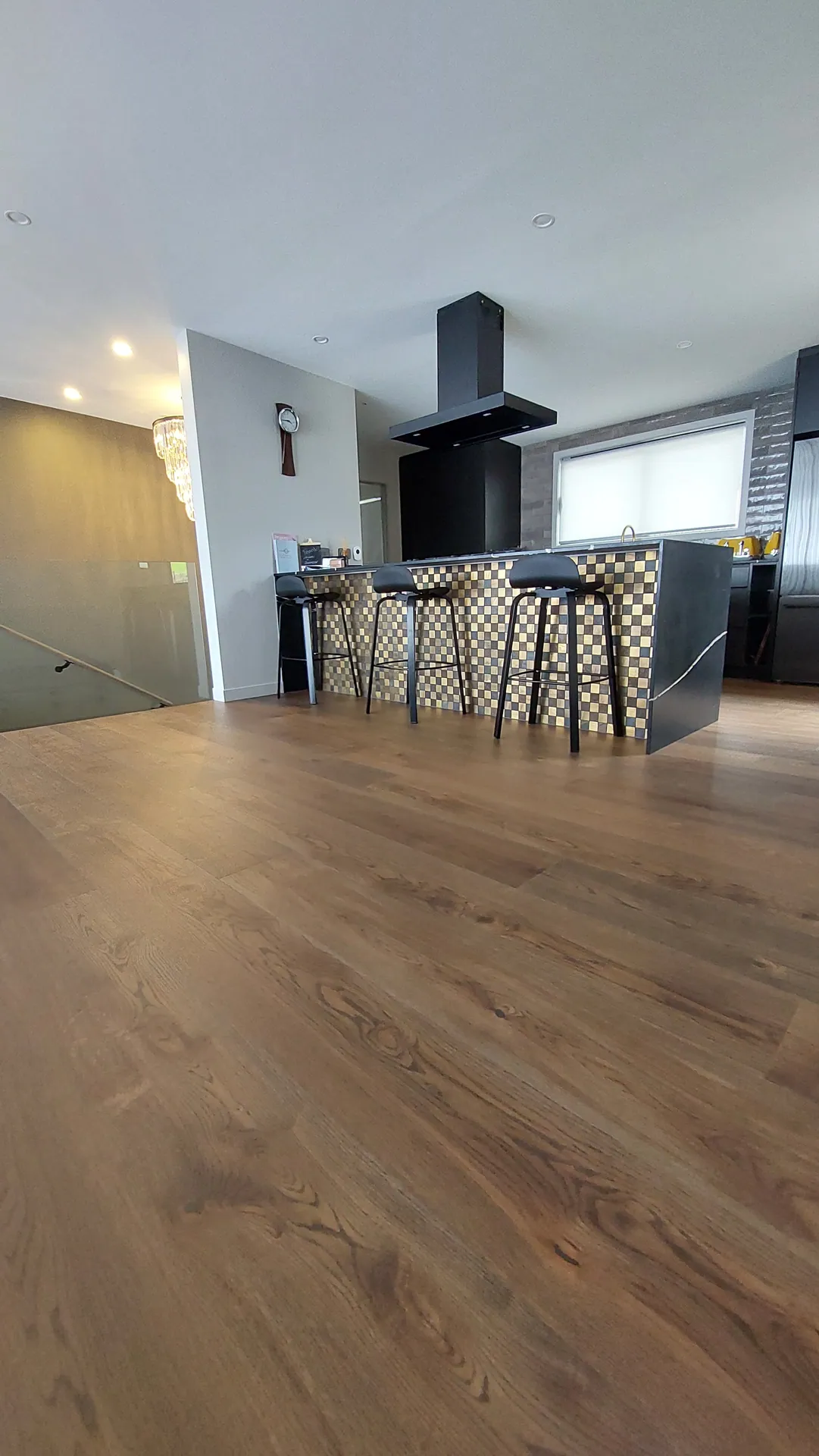 A wide brown oak floor in front of a kitchen island with a patterned front, black bar stools and a black range hood.