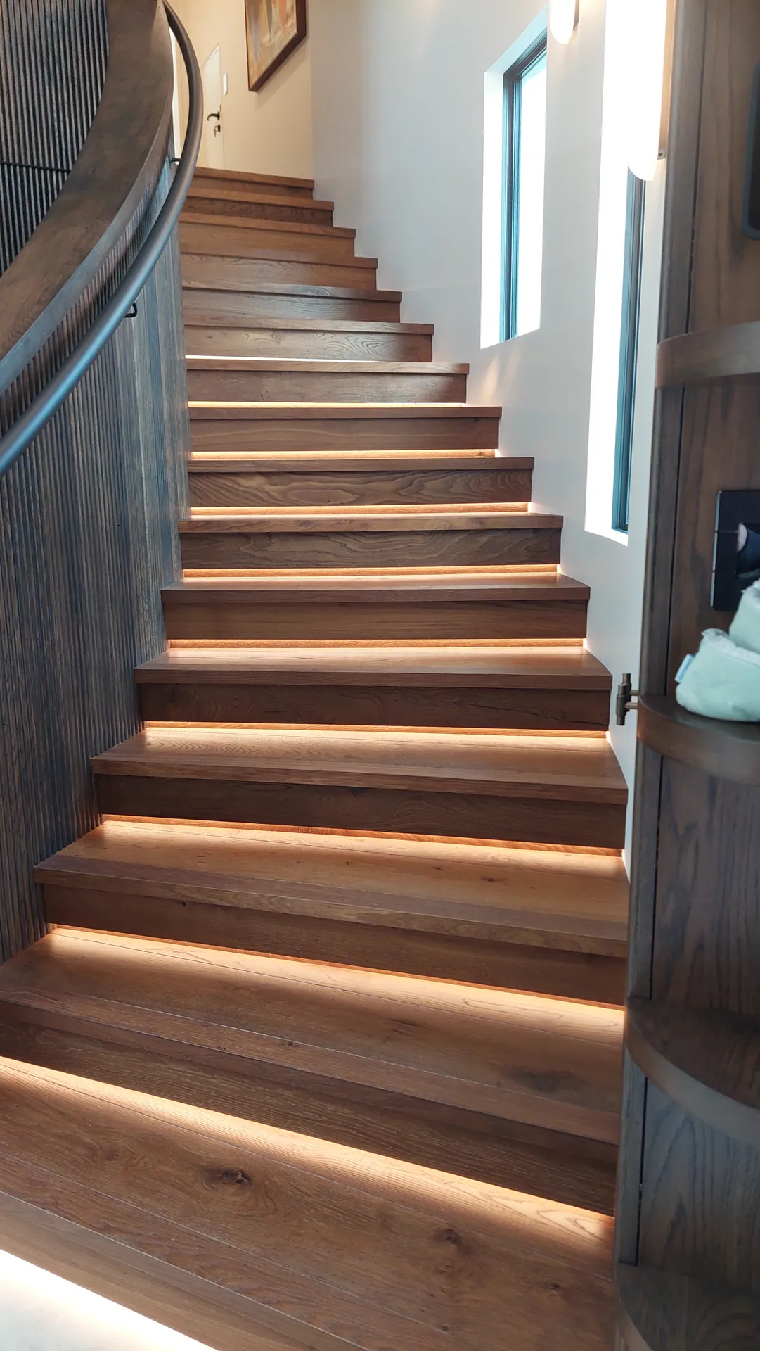A curved staircase with oak treads and risers, each tread lit underneath by a warm strip light.