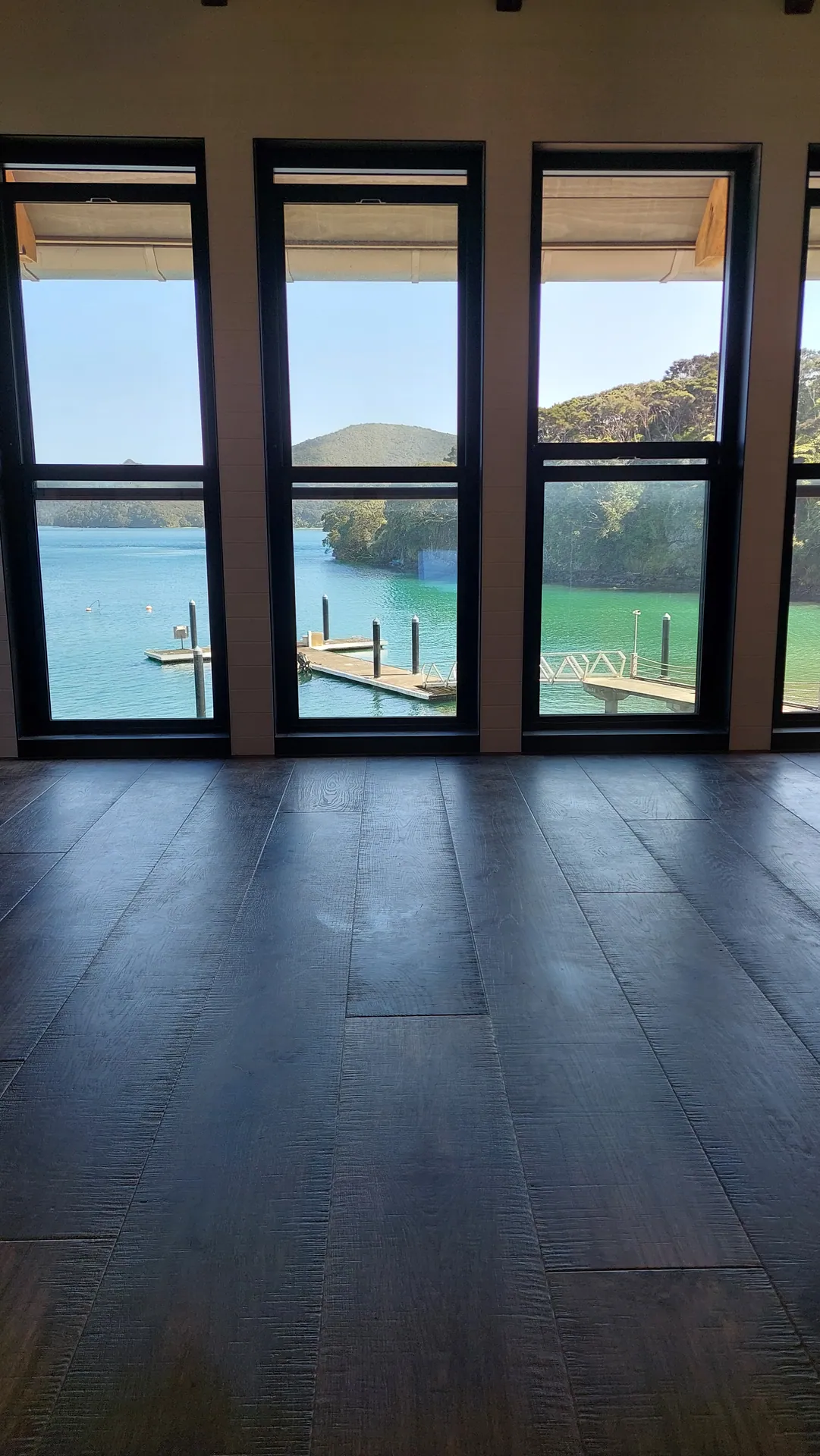 A dark wide-board floor in front of tall windows looking out over a harbour, jetty and bush-covered hills.