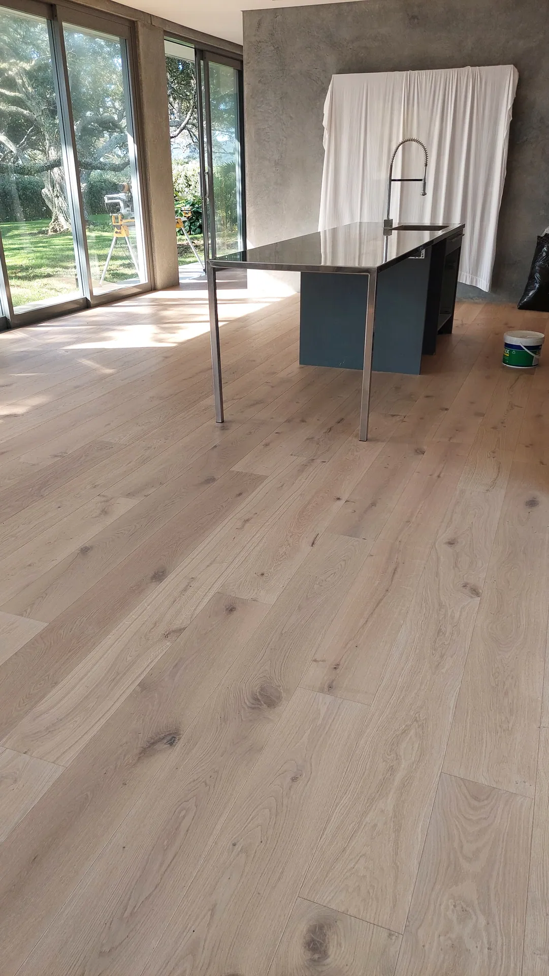 A finished light oak floor with knots running past a stainless steel kitchen island and glass doors to a garden.