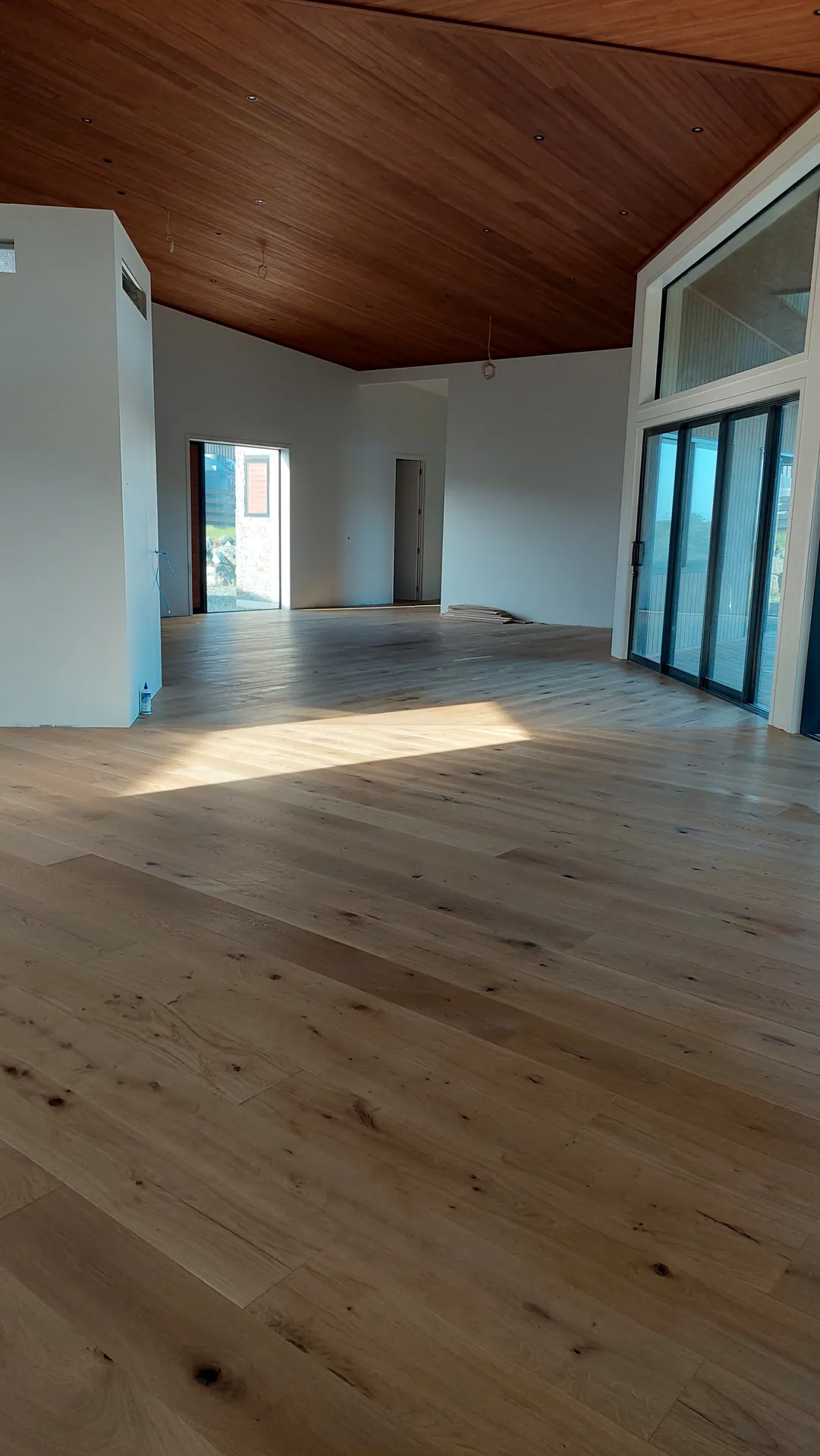 An empty open room with a knotty light oak floor, a sunlit patch near the glass doors and a timber-lined ceiling.