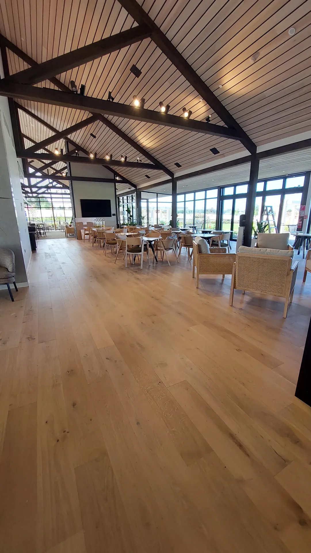 A large dining hall with exposed timber trusses, rows of chairs and tables, and a light oak floor running to a wall of windows.