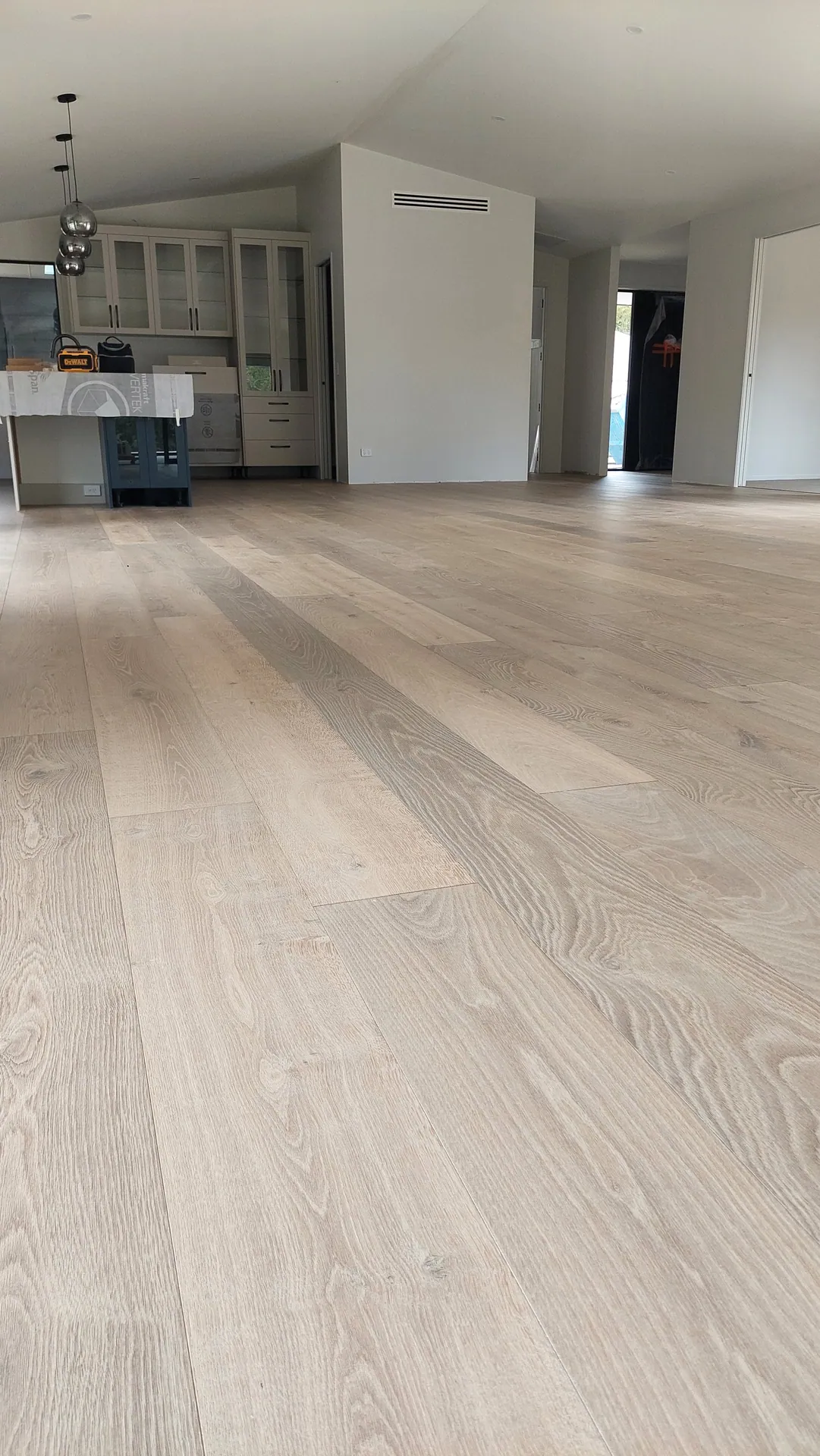 A low close view along pale oak boards with visible grain toward a kitchen with glass cabinets.