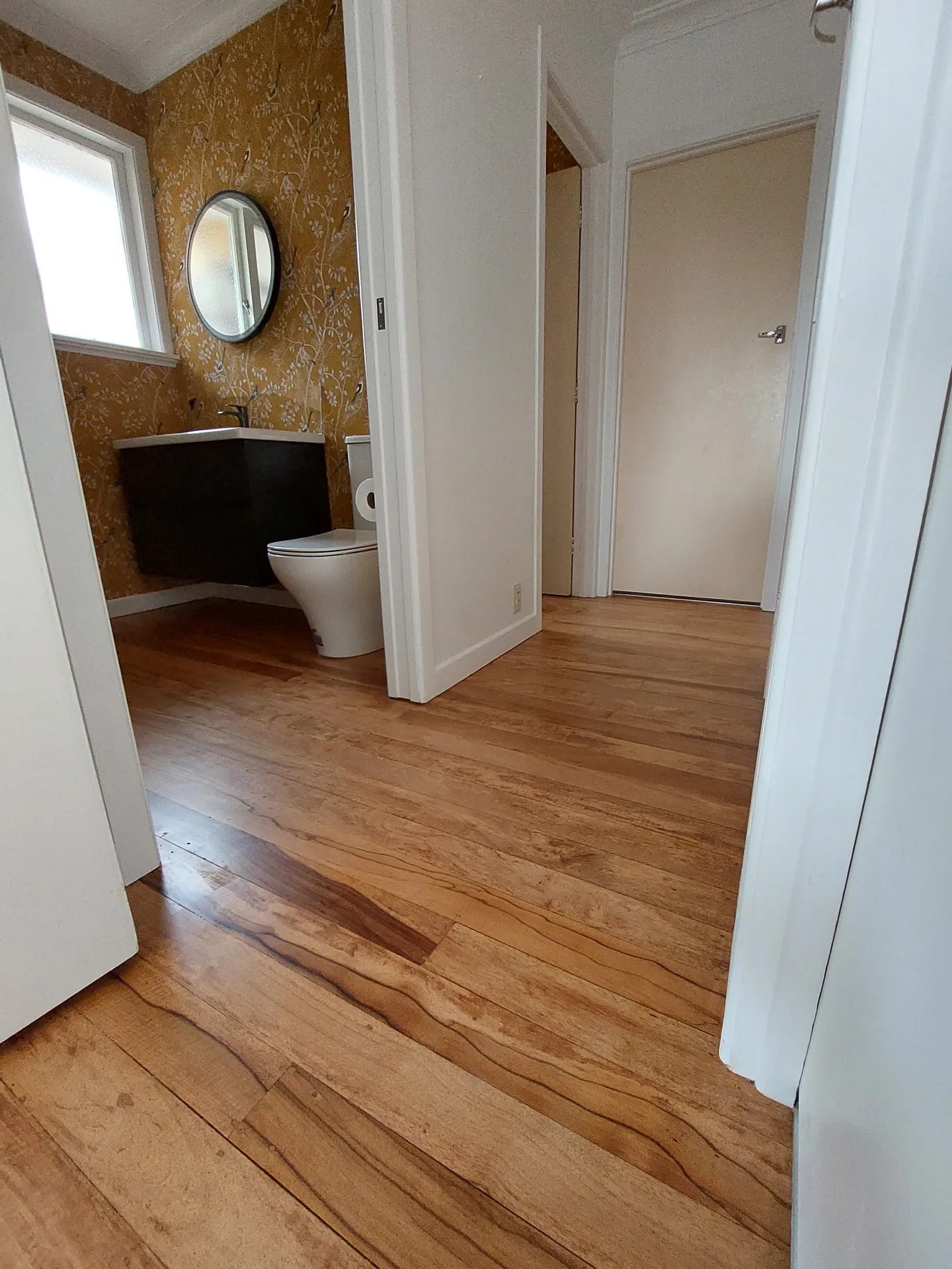Refinished native timber floorboards running from a hallway into a bathroom with a round mirror.