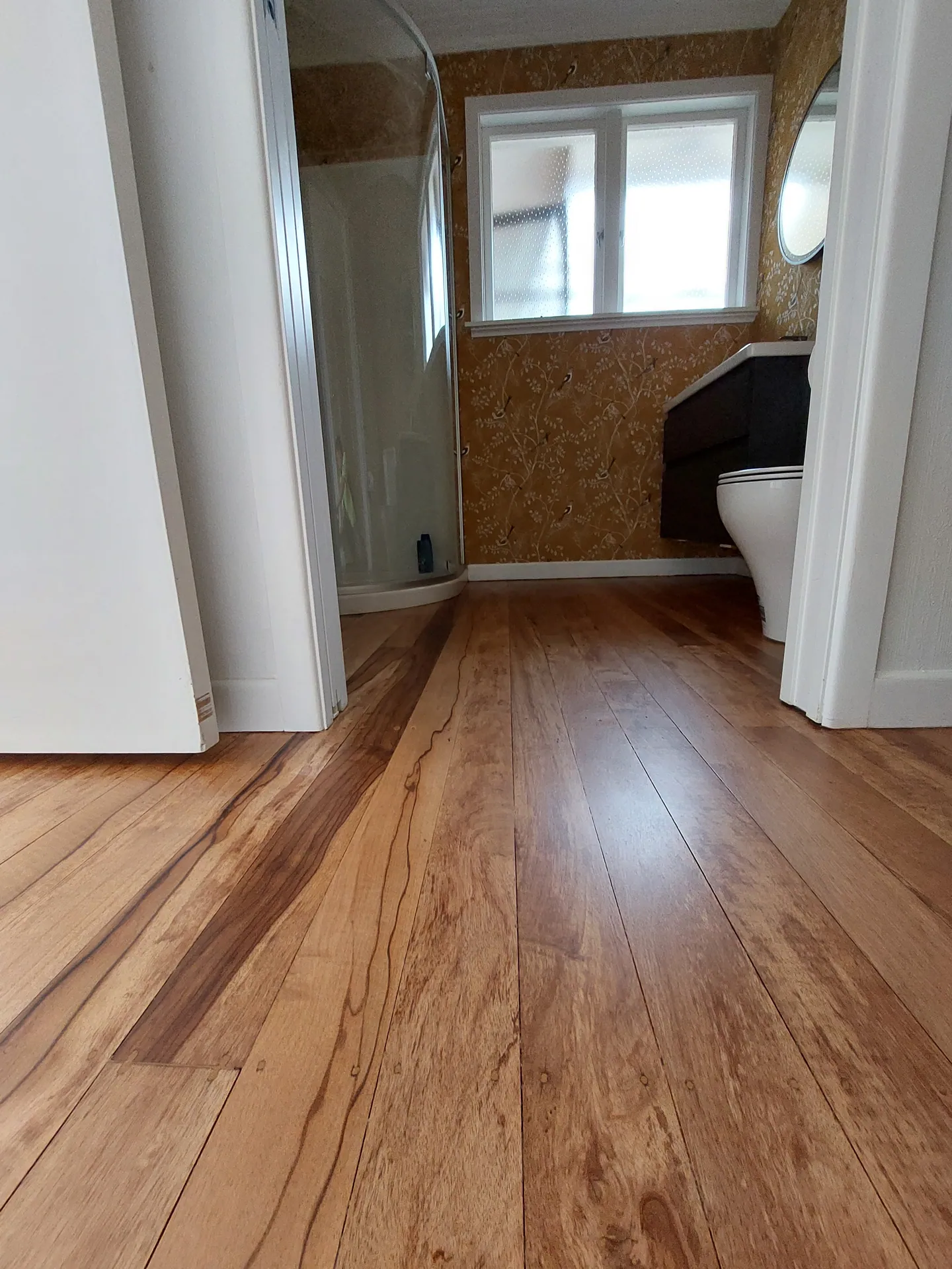 Refinished native timber floorboards in a bathroom with gold bird wallpaper, a shower and a vanity.