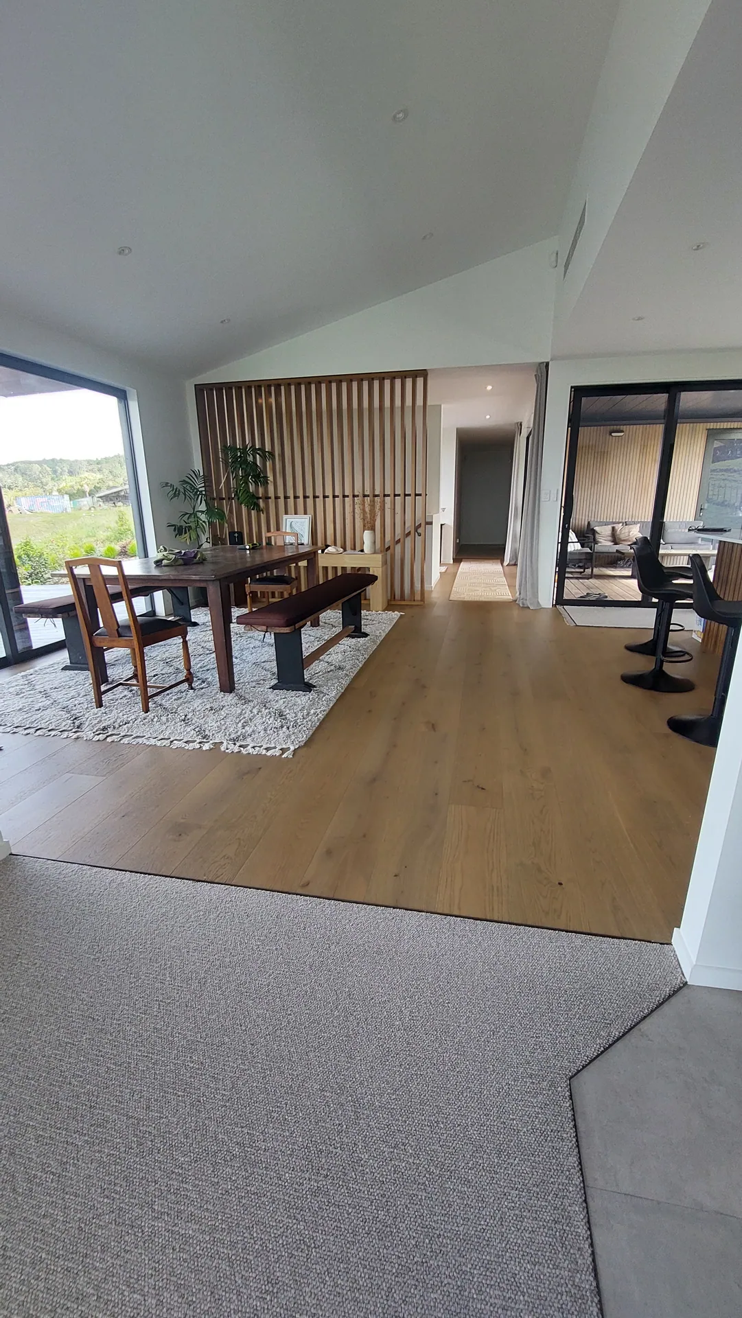 A dining area with an oak floor, a timber batten screen and a long table, with carpet in the foreground.