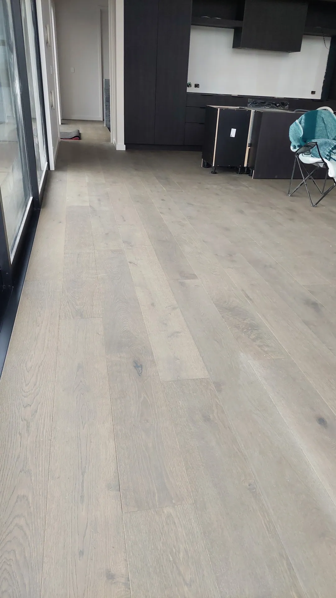Pale limed oak boards running along a wall of glass doors toward a dark kitchen still being installed.