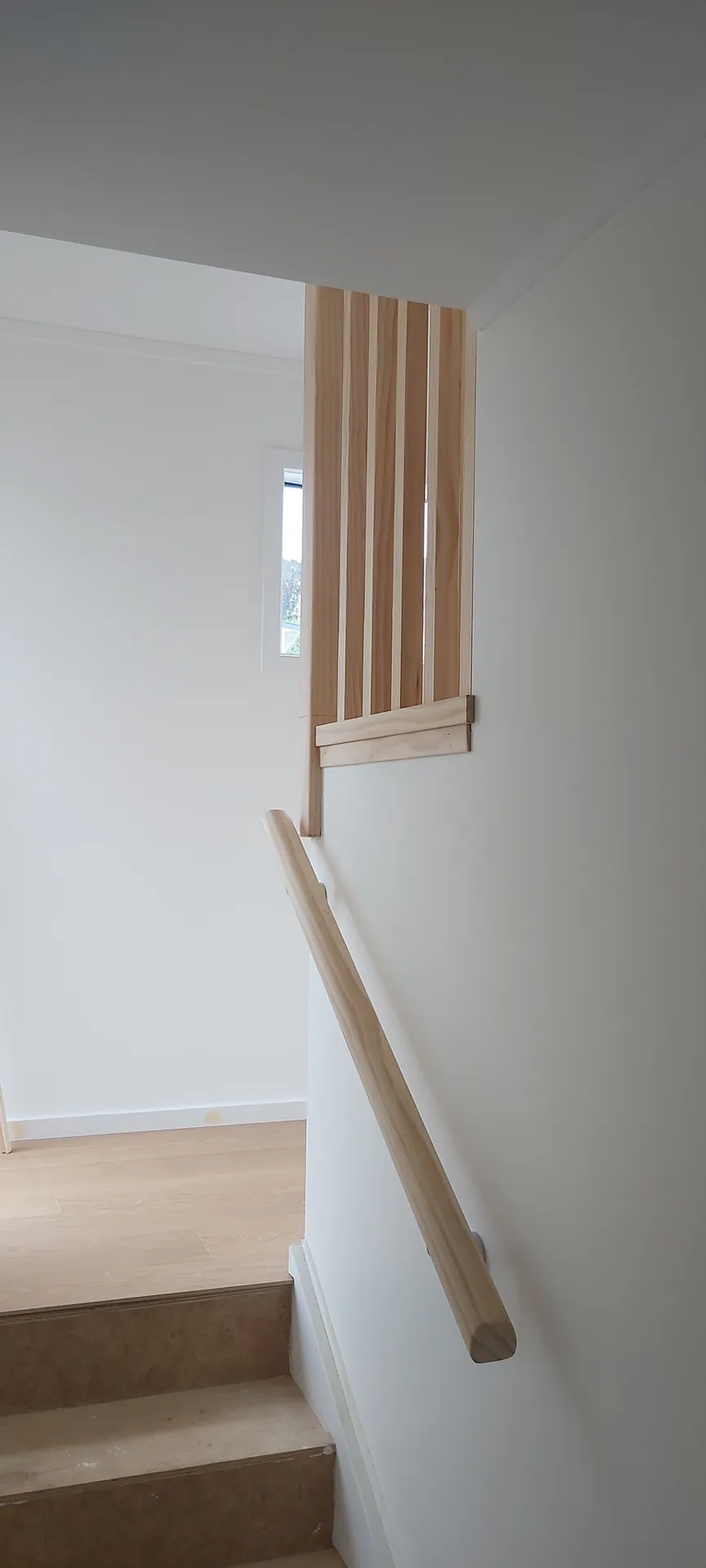 Pine handrail fixed to a white wall beside plywood stair treads, with a slatted pine screen above.