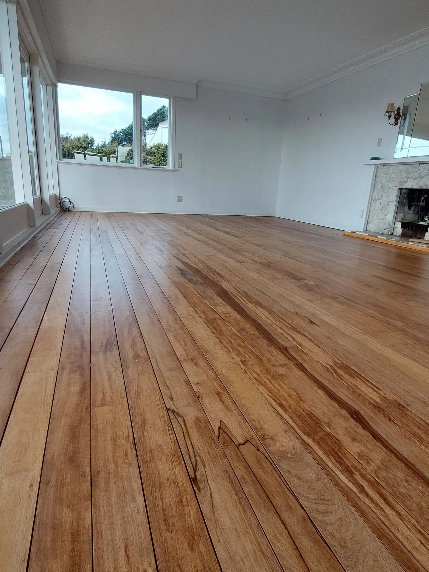 Low-angle view across refinished native timber floorboards in a lounge towards windows and a fireplace.