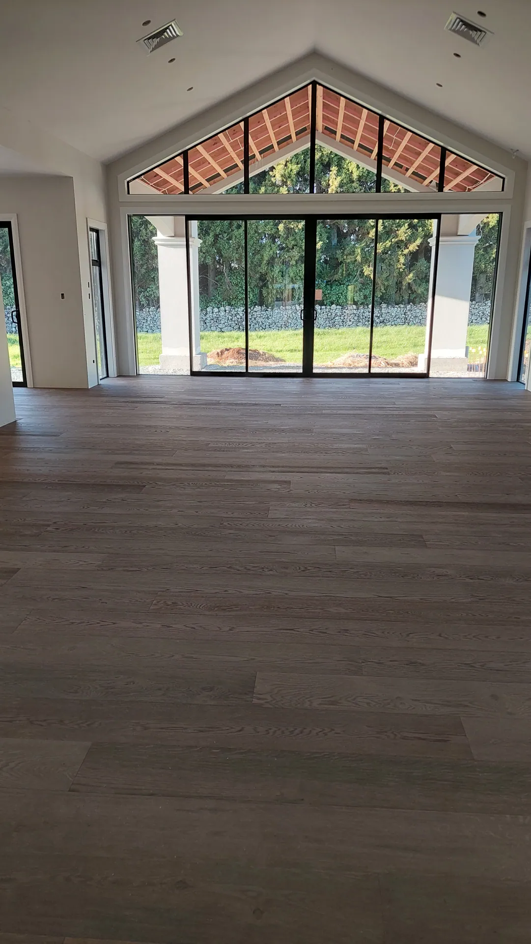 A large empty living room with a dark smoked oak floor facing glass doors under a tall gable window.