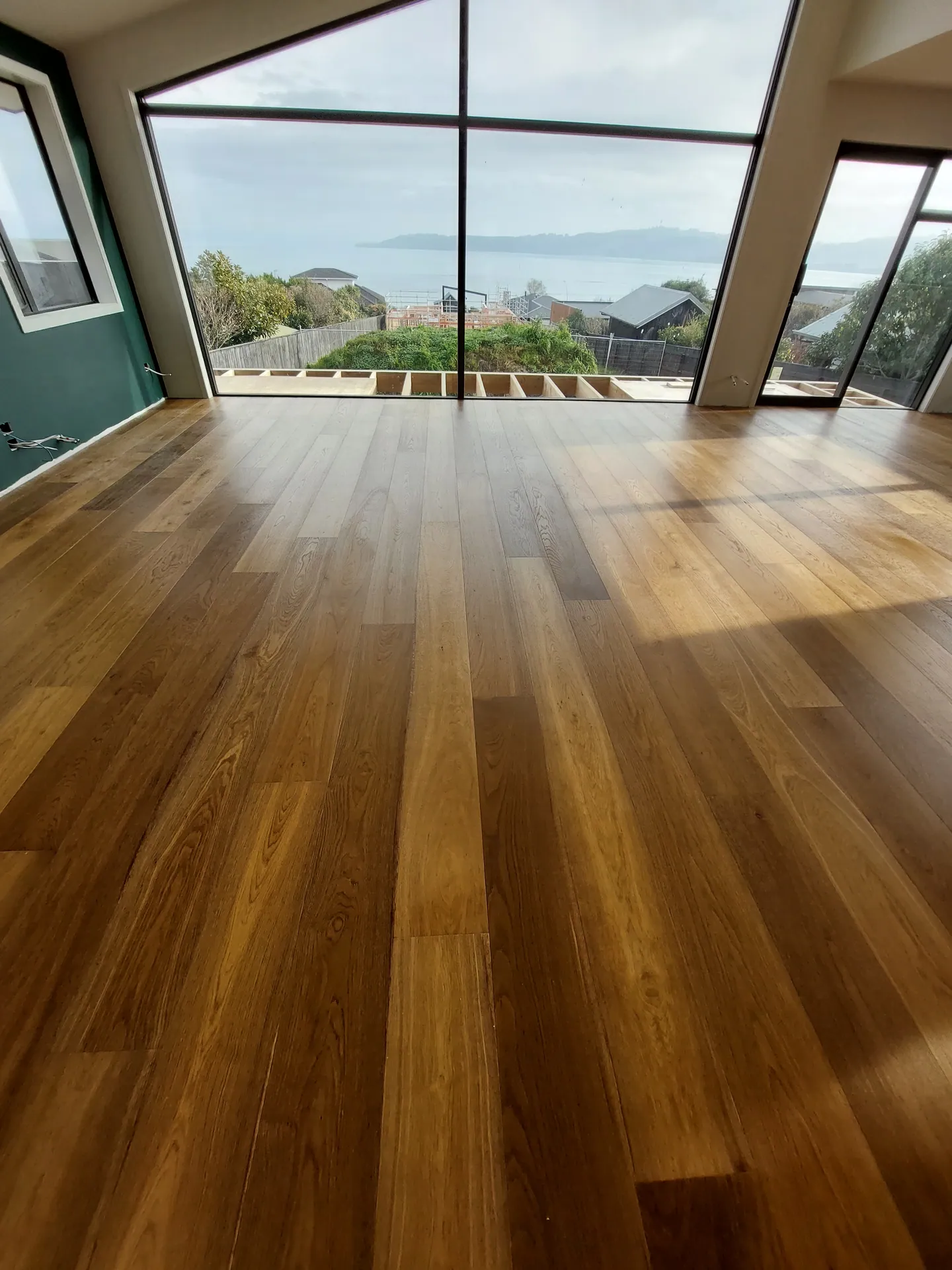 Rich brown timber floorboards catching sunlight in front of a large raked window looking out to the sea.