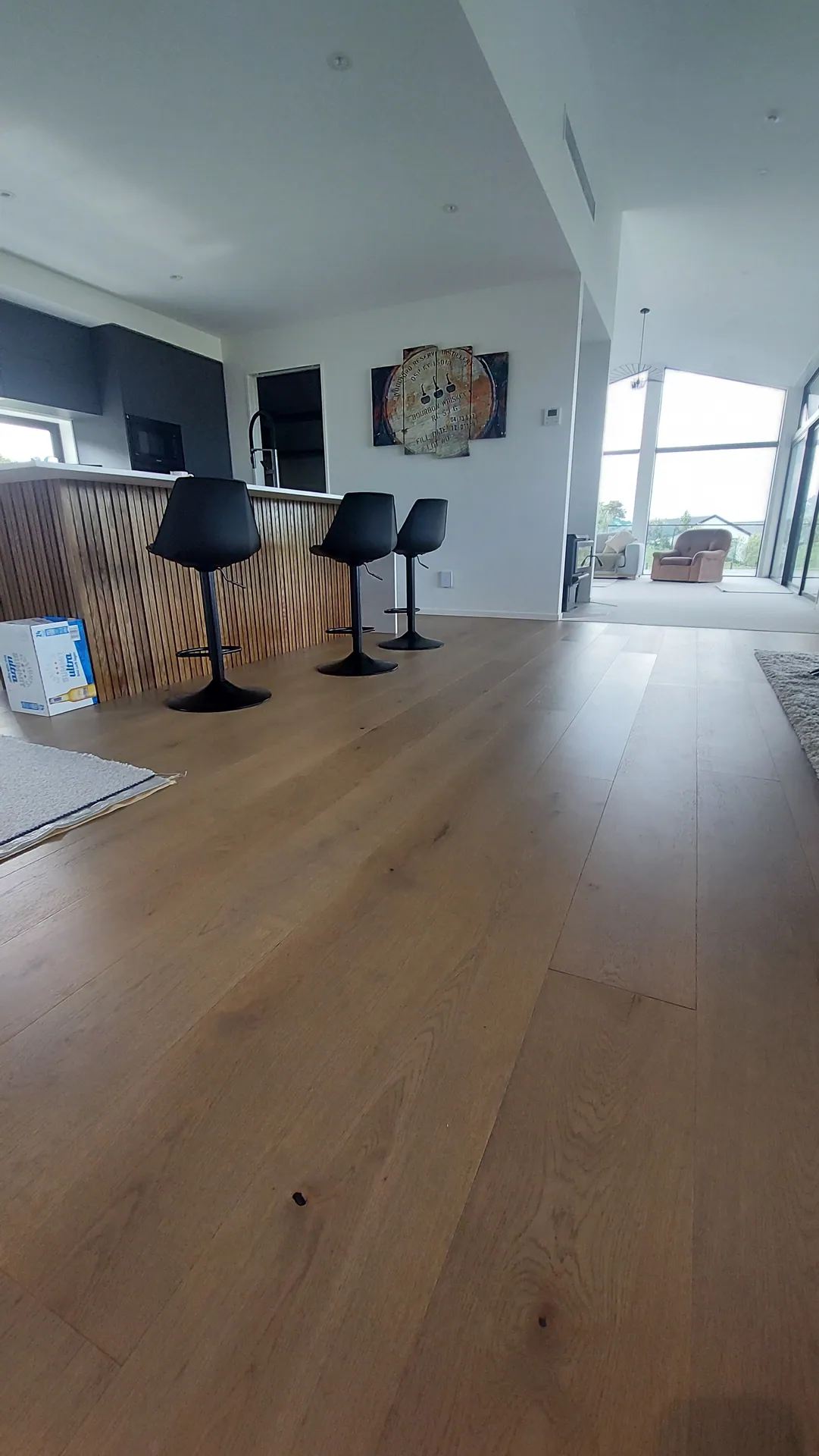 A wide oak floor in front of a timber-batten kitchen island with black bar stools.