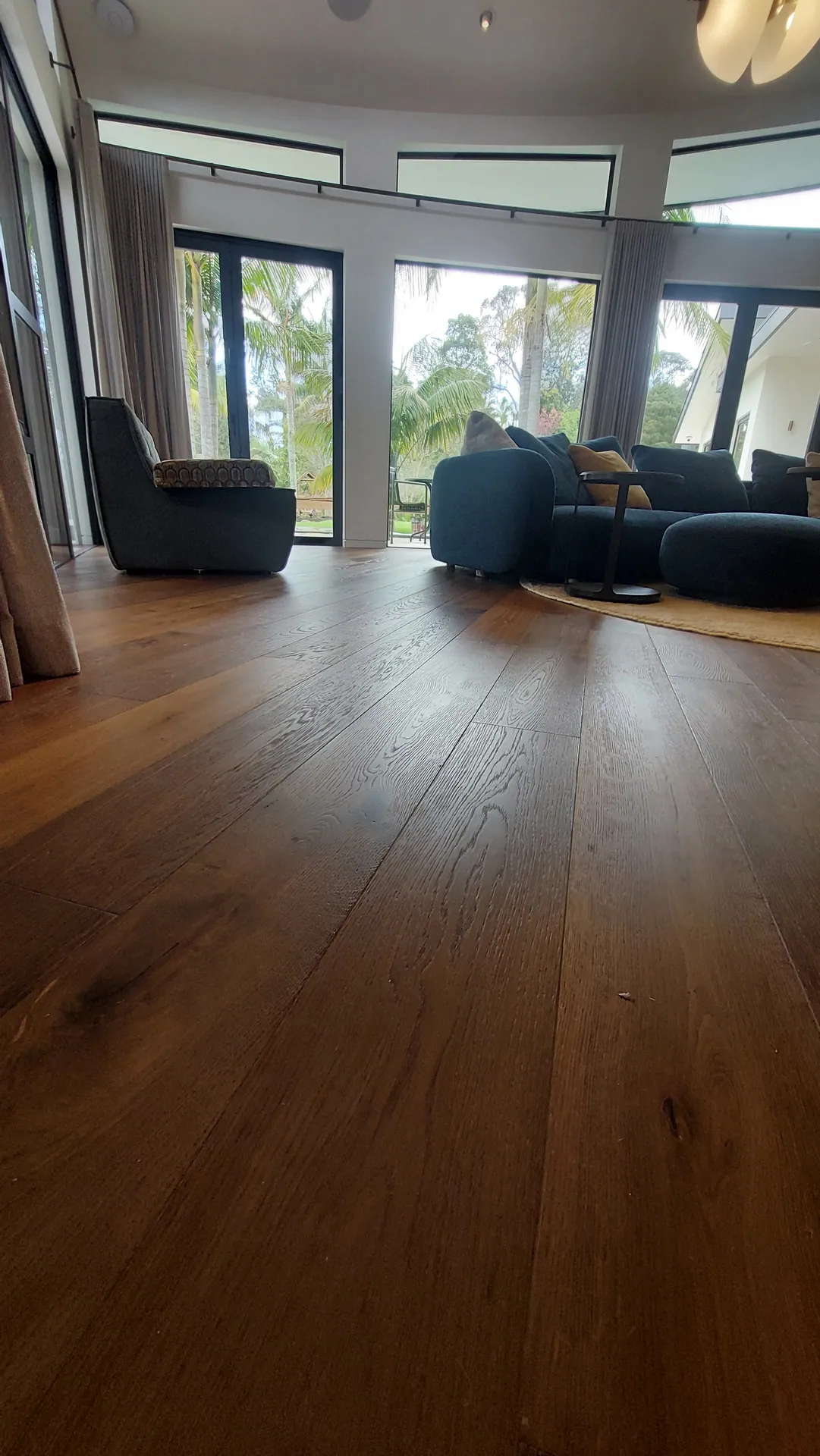 A low view across wide brown oak boards in a living room with dark sofas and tall curved windows.