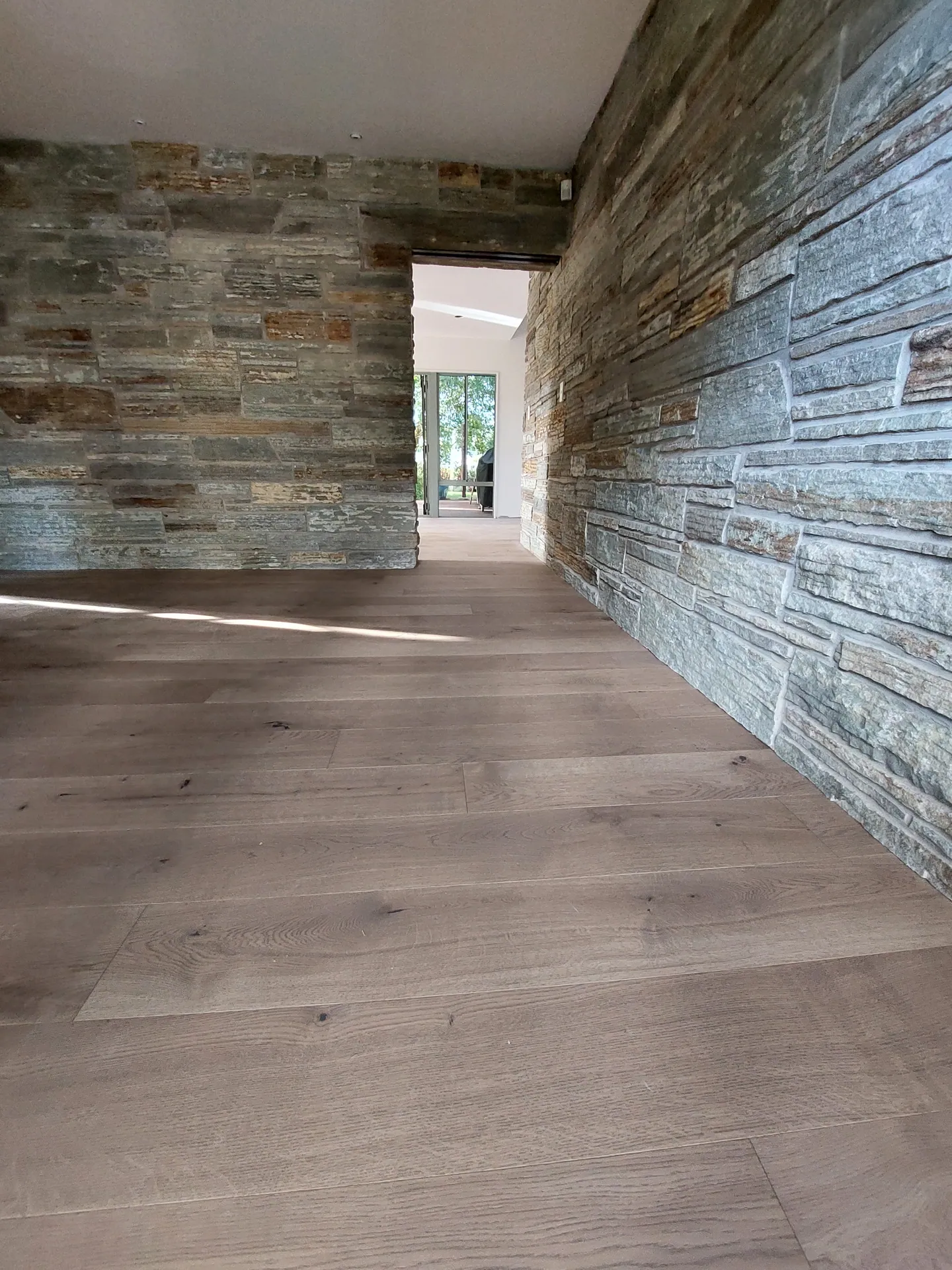 Wide grey-toned timber floorboards running alongside a stacked stone feature wall towards a doorway.